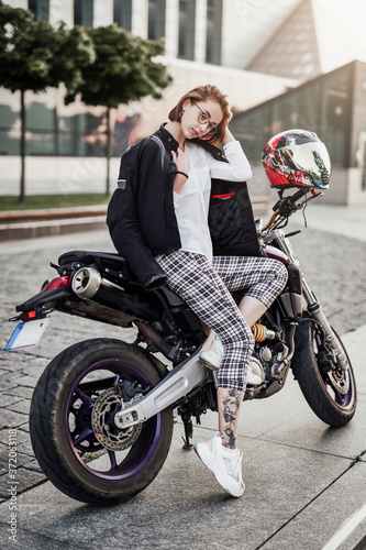 Cute and stylish looking girl is sitting on her purple motorcycle. She wears a black motorcycle jacket and plaid pants.