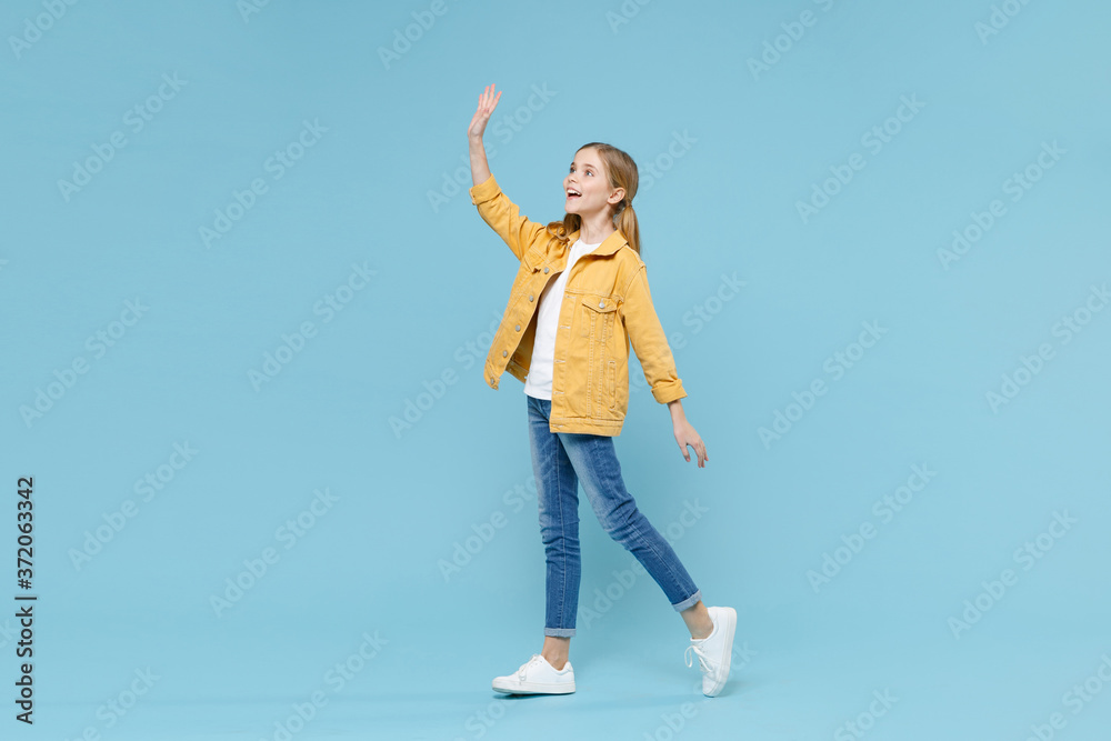 Full length portrait of funny little blonde kid girl 12-13 years old in yellow jacket isolated on blue background studio. Childhood lifestyle concept. Waving and greeting with hand as notices someone. Full length portrait of funny little blonde kid girl 12-13 years old in yellow jacket isolated on blue background studio. Childhood lifestyle concept. Waving and greeting with hand as notices someone.