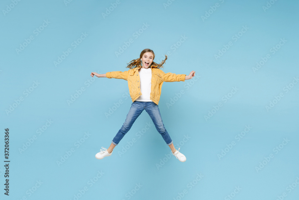 Full length portrait of excited little blonde kid girl 12-13 years old in yellow jacket isolated on blue background studio. Childhood lifestyle concept. Mock up copy space. Jumping, spreading hands.