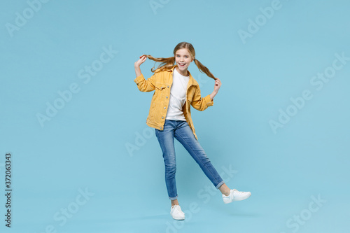 Full length portrait of funny little blonde kid girl 12-13 years old in yellow jacket posing isolated on blue wall background studio. Childhood lifestyle concept. Mock up copy space. Hold ponytails.