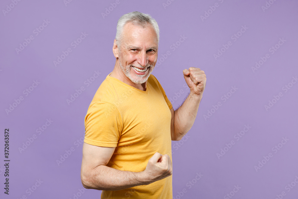 Joyful elderly gray-haired mustache bearded man in casual yellow t-shirt posing isolated on pastel violet background studio. People lifestyle concept. Mock up copy space. Clenching fists like winner. Joyful elderly gray-haired mustache bearded man in casual yellow t-shirt posing isolated on pastel violet background studio. People lifestyle concept. Mock up copy space. Clenching fists like winner.
