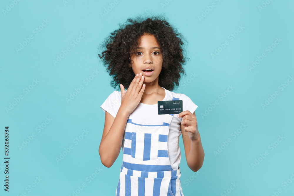 Shocked little african american kid girl 12-13 years old in striped clothes isolated on blue background. Childhood lifestyle concept. Mock up copy space Hold credit bank card covering mouth with hand. Shocked little african american kid girl 12-13 years old in striped clothes isolated on blue background. Childhood lifestyle concept. Mock up copy space Hold credit bank card covering mouth with hand.