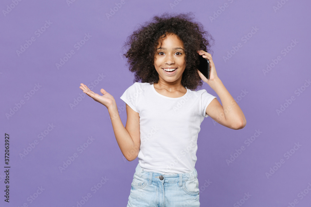 Excited little african american kid girl 12-13 years old in white t-shirt isolated on violet background studio. Childhood lifestyle concept. Mock up copy space. Talk on mobile phone, spreading hands. Excited little african american kid girl 12-13 years old in white t-shirt isolated on violet background studio. Childhood lifestyle concept. Mock up copy space. Talk on mobile phone, spreading hands.
