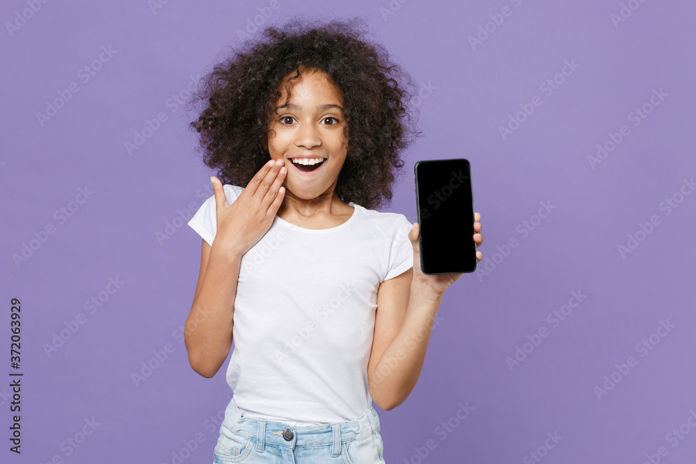 Excited little african american kid girl 12-13 years old in white t-shirt isolated on violet wall background. Childhood lifestyle concept. Hold mobile phone with blank empty screen, put hand on cheek. Excited little african american kid girl 12-13 years old in white t-shirt isolated on violet wall background. Childhood lifestyle concept. Hold mobile phone with blank empty screen, put hand on cheek.
