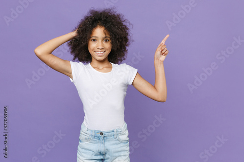 Smiling little african american kid girl 12-13 years old in white t-shirt isolated on violet wall background studio portrait. Childhood lifestyle concept. Pointing index finger up, put hand on head.