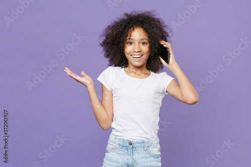 Excited little african american kid girl 12-13 years old in white t-shirt isolated on violet background studio. Childhood lifestyle concept. Mock up copy space. Talk on mobile phone, spreading hands.