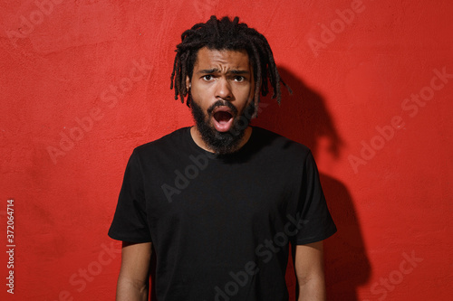 Shocked worried young african american man guy with dreadlocks 20s wearing black casual t-shirt posing keeping mouth open looking camera isolated on bright red color wall background studio portrait.