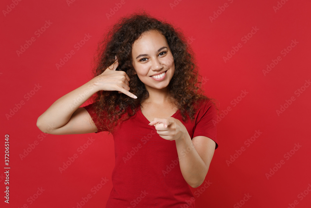 Smiling african american girl in casual t-shirt isolated on red background. People lifestyle concept. Mock up copy space. Doing phone gesture like says call me back, pointing index finger on camera. Smiling african american girl in casual t-shirt isolated on red background. People lifestyle concept. Mock up copy space. Doing phone gesture like says call me back, pointing index finger on camera.