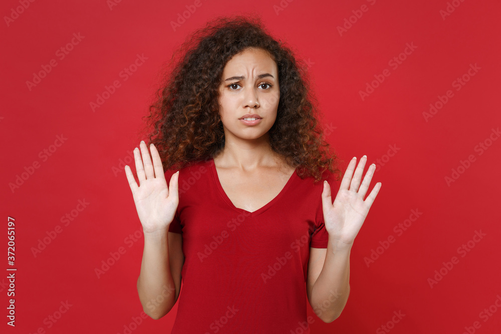 Concerned young african american woman girl in casual t-shirt posing isolated on red wall background studio portrait. People emotions lifestyle concept. Mock up copy space. Rising hands showing palms. Concerned young african american woman girl in casual t-shirt posing isolated on red wall background studio portrait. People emotions lifestyle concept. Mock up copy space. Rising hands showing palms.