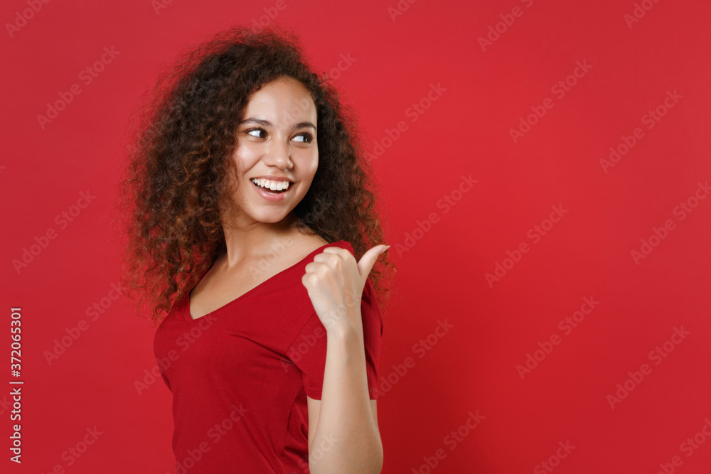 Side view of smiling young african american woman girl in casual t-shirt posing isolated on red wall background studio portrait. People lifestyle concept. Mock up copy space. Pointing thumb aside. Side view of smiling young african american woman girl in casual t-shirt posing isolated on red wall background studio portrait. People lifestyle concept. Mock up copy space. Pointing thumb aside.
