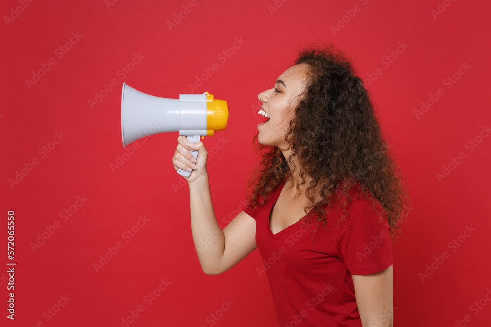 Side view of funny young african american woman girl in casual t-shirt posing isolated on red background studio portrait. People emotions lifestyle concept. Mock up copy space. Screaming in megaphone. Side view of funny young african american woman girl in casual t-shirt posing isolated on red background studio portrait. People emotions lifestyle concept. Mock up copy space. Screaming in megaphone.