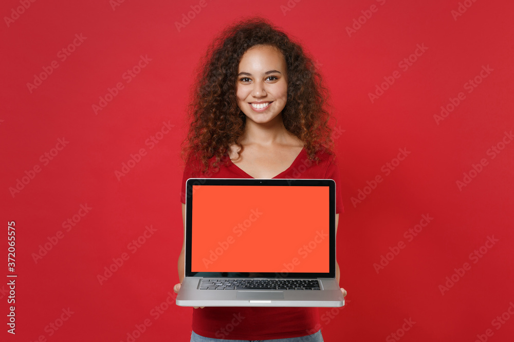 Smiling young african american woman girl in casual t-shirt posing isolated on red background studio. People lifestyle concept. Mock up copy space. Hold laptop pc computer with blank empty screen. Smiling young african american woman girl in casual t-shirt posing isolated on red background studio. People lifestyle concept. Mock up copy space. Hold laptop pc computer with blank empty screen.
