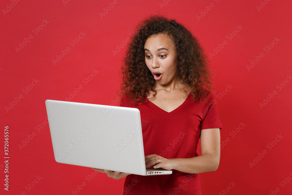 Naklejka premium Shocked young african american woman girl in casual t-shirt posing isolated on red background studio portrait. People emotions lifestyle concept. Mock up copy space. Working on laptop pc computer.