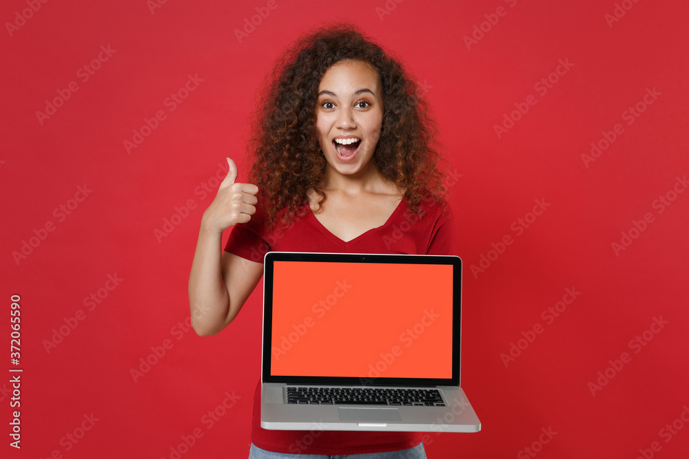Naklejka premium Excited young african american woman girl in casual t-shirt isolated on red background. People lifestyle concept. Mock up copy space. Hold laptop pc computer with blank empty screen, showing thumb up.