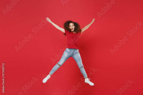 Full length portrait of cheerful young african american girl in casual t-shirt isolated on red background studio portrait. People lifestyle concept. Mock up copy space. Jumping spreading hands legs.