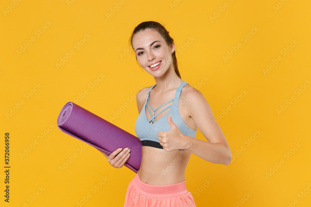 Smiling young fitness sporty woman girl in sportswear posing working out isolated on yellow background studio. Workout sport motivation concept. Mock up copy space. Hold yoga mat, showing thumb up.