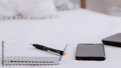 Primer plano de anotador junto a un lápiz y un teléfono móvil sobre una cama de blancas sábanas en una habitación blanca e iluminada. Concepto de trabajar desde el hogar. 