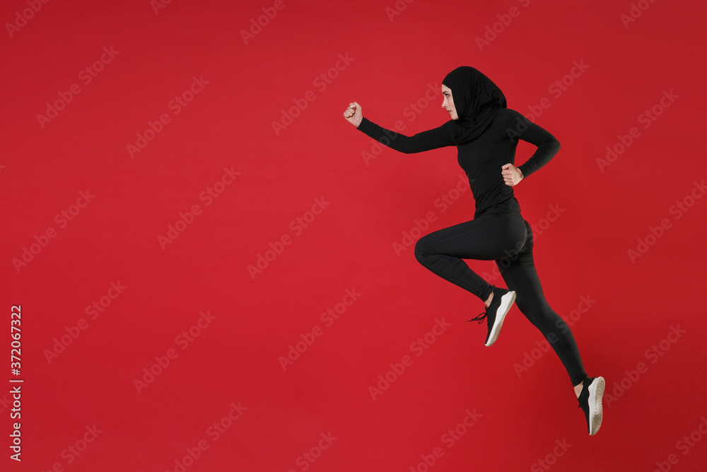 Full length portrait side view of young arabian muslim woman in hijab black clothes posing isolated on red wall background studio portrait. People religious lifestyle concept. Jumping like running.