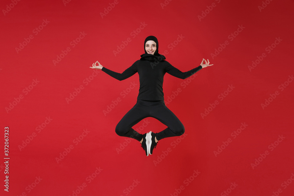 Fototapeta Full length portrait of smiling young arabian muslim woman in hijab black clothes posing isolated on red background studio. People religious lifestyle concept. Jumping, hold hands in yoga gesture.