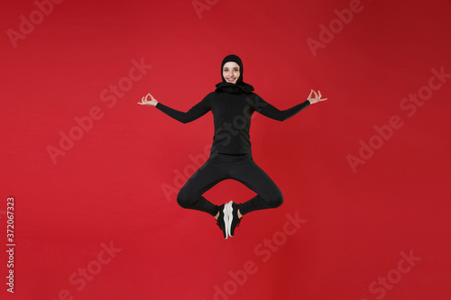Full length portrait of smiling young arabian muslim woman in hijab black clothes posing isolated on red background studio. People religious lifestyle concept. Jumping, hold hands in yoga gesture.