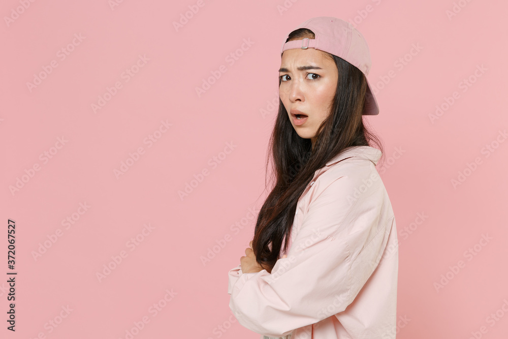 Side view of shocked worried young asian woman girl in casual clothes cap posing isolated on pastel pink background. People lifestyle concept. Mock up copy space. Holding hands crossed looking camera.