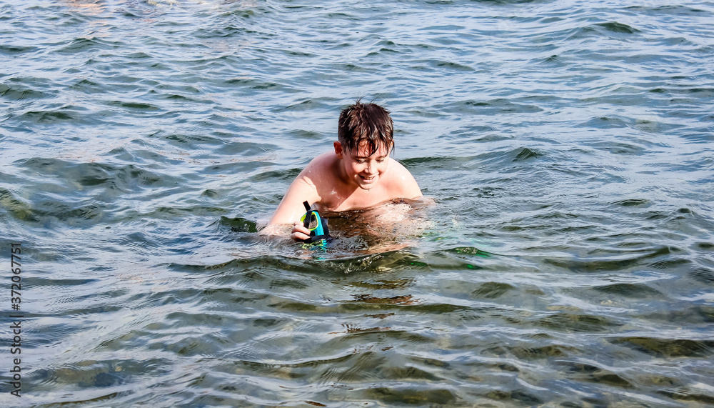 A boy swims in the lake in the summer, holding a diving mask in his hand.