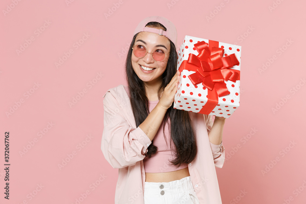 Pensive young asian woman in casual clothes cap glasses isolated on pastel pink wall background. St. Valentine's Day, Women's Day, birthday, holiday concept. Hold red present box with gift ribbon bow. Pensive young asian woman in casual clothes cap glasses isolated on pastel pink wall background. St. Valentine's Day, Women's Day, birthday, holiday concept. Hold red present box with gift ribbon bow.