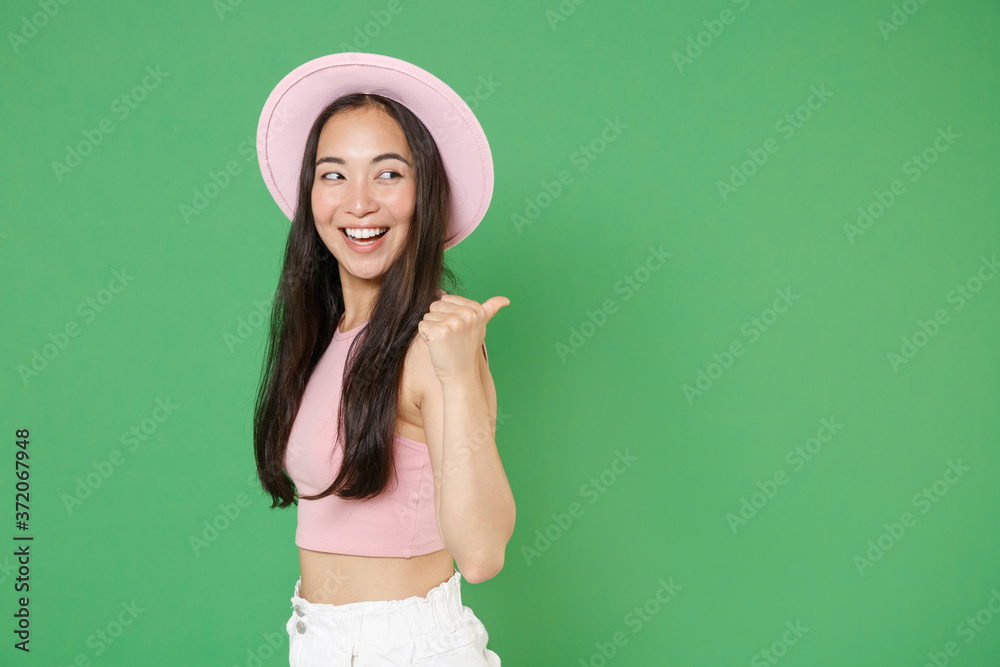 Side view of smiling young asian woman girl in casual pink clothes hat posing isolated on green background studio portrait. People emotions lifestyle concept. Mock up copy space. Pointing thumb aside. Side view of smiling young asian woman girl in casual pink clothes hat posing isolated on green background studio portrait. People emotions lifestyle concept. Mock up copy space. Pointing thumb aside.