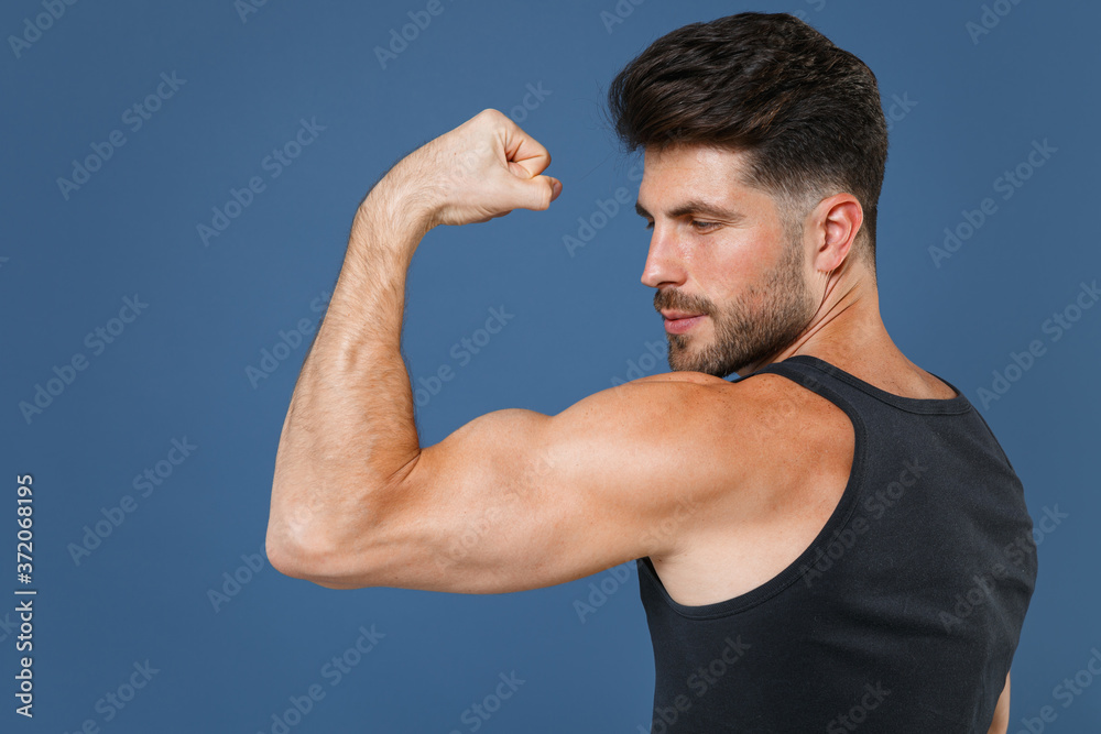 Side view of strong young bearded fitness sporty guy sportsman in black undershirt isolated on blue wall background studio portrait. Workout sport motivation lifestyle concept. Showing biceps muscles. Side view of strong young bearded fitness sporty guy sportsman in black undershirt isolated on blue wall background studio portrait. Workout sport motivation lifestyle concept. Showing biceps muscles.