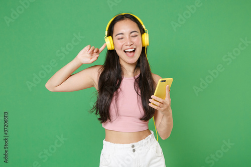 Cheerful young asian woman girl in pink clothes isolated on green background studio. People lifestyle concept. Mock up copy space. Listening music with headphones, mobile phone, sing song dancing. photo