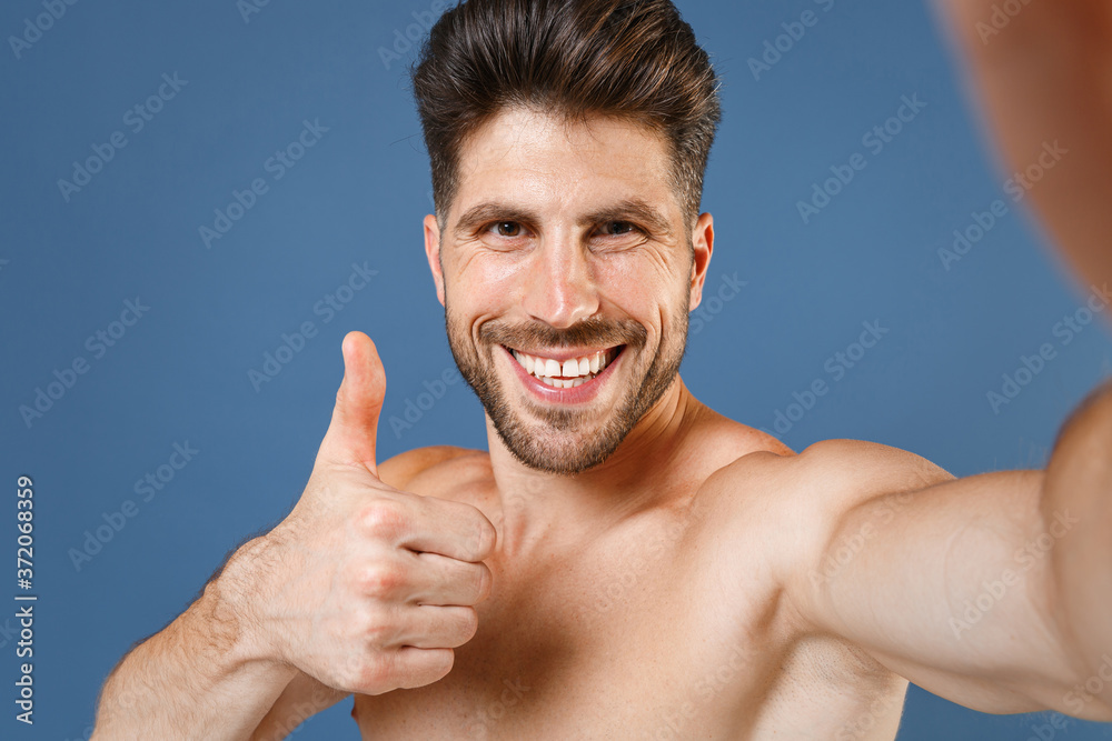 Close up smiling fitness sporty strong guy bare-chested muscular sportsman isolated on blue background. Workout sport motivation lifestyle concept. Doing selfie shot on mobile phone, showing thumb up. Close up smiling fitness sporty strong guy bare-chested muscular sportsman isolated on blue background. Workout sport motivation lifestyle concept. Doing selfie shot on mobile phone, showing thumb up.