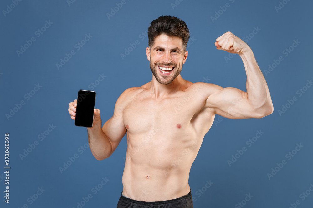 Funny young fitness sporty guy bare-chested sportsman isolated on blue background. Workout sport motivation lifestyle concept. Mock up copy space. Hold mobile phone with blank screen showing biceps. Funny young fitness sporty guy bare-chested sportsman isolated on blue background. Workout sport motivation lifestyle concept. Mock up copy space. Hold mobile phone with blank screen showing biceps.