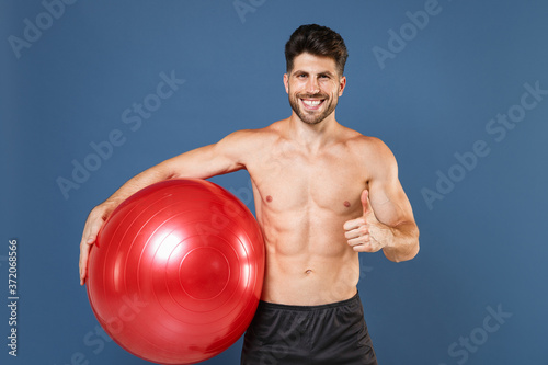 Smiling young bearded fitness sporty strong guy bare-chested muscular sportsman isolated on blue background studio portrait. Workout sport motivation lifestyle concept. Hold fitball, showing thumb up.