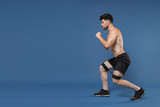 Full length portrait side view of fitness sporty guy bare-chested muscular sportsman isolated on blue background. Workout sport motivation lifestyle concept. Doing exercises lunge with fitness gums.