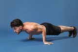 Full length portrait of young fitness sporty strong guy bare-chested muscular sportsman isolated on blue background. Workout sport motivation lifestyle concept. Doing push-ups exercises looking aside.