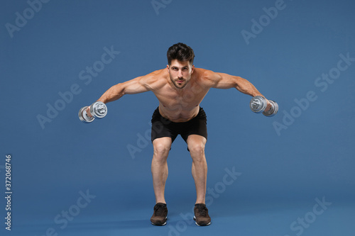 Full length portrait of young bearded fitness sporty strong guy bare-chested muscular sportsman isolated on blue background. Workout sport motivation lifestyle concept. Doing exercise with dumbbells. © ViDi Studio