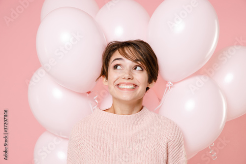 Smiling pensive pretty young brunette woman girl in knitted casual sweater celebrating posing on pastel pink background with air baloons. Birthday holiday party, people emotions concept. Looking up.