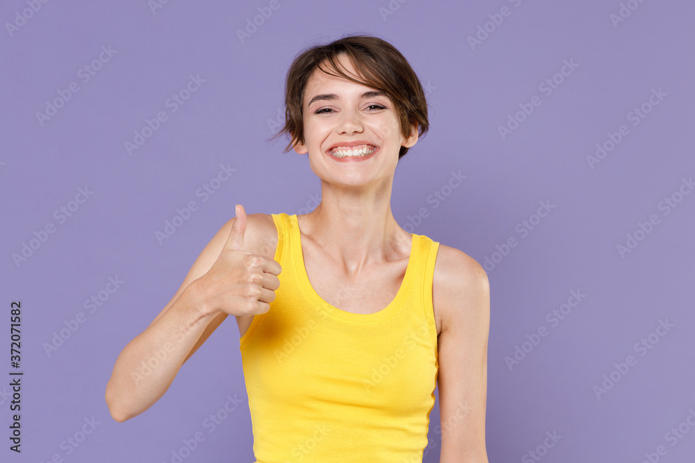 Smiling funny young brunette woman girl wearing yellow casual tank top posing isolated on pastel violet wall background studio portrait. People sincere emotions lifestyle concept. Showing thumb up. Smiling funny young brunette woman girl wearing yellow casual tank top posing isolated on pastel violet wall background studio portrait. People sincere emotions lifestyle concept. Showing thumb up.