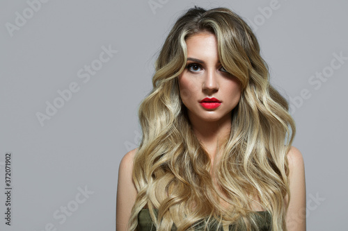 Blonde with curls on a gray background. Long hair. Red lipstick. Beautiful lips and big eyes. Portrait