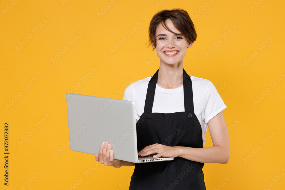Smiling young female woman 20s barista bartender barman employee in white t-shirt apron posing working on laptop pc computer looking camera isolated on yellow color wall background studio portrait. Smiling young female woman 20s barista bartender barman employee in white t-shirt apron posing working on laptop pc computer looking camera isolated on yellow color wall background studio portrait.