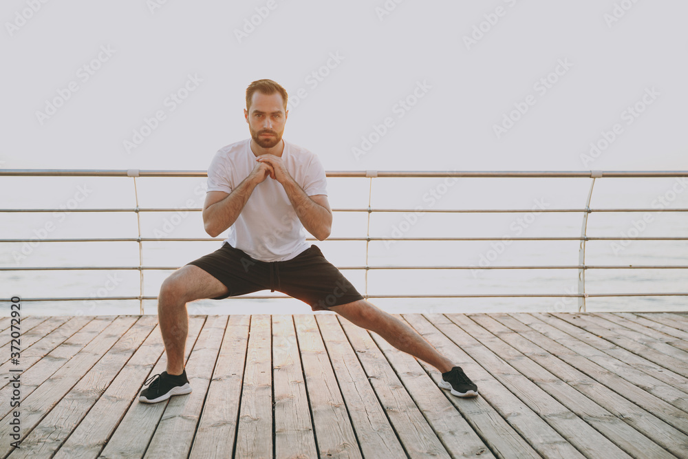 Full length portrait of handsome young bearded athletic man guy 20s in white t-shirt posing training doing stretching lunge exercising for legs looking camera at sunrise over the sea outdoors. - obrazy, fototapety, plakaty 