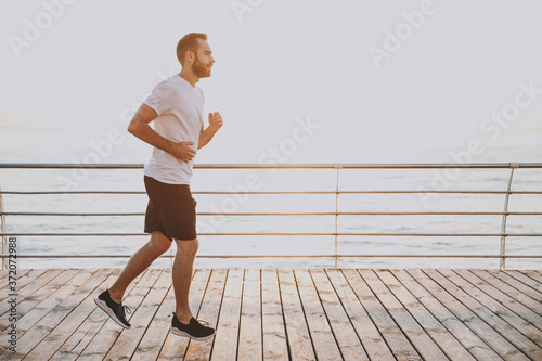 Side view full length portrait of handsome attractive young bearded athletic man guy 20s in casual white t-shirt black shorts posing training running looking aside at sunrise over the sea outdoors.