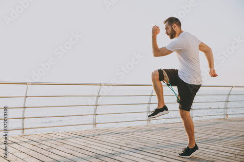 Full length portrait handsome attractive young bearded athletic man guy 20s in white t-shirt posing training doing exercises for legs with fitness gums looking aside at sunrise over the sea outdoors.