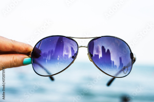 Female hand holds colorful sunglasses against the background of the sky and blue sea. The points reflect the storey buildings of the city