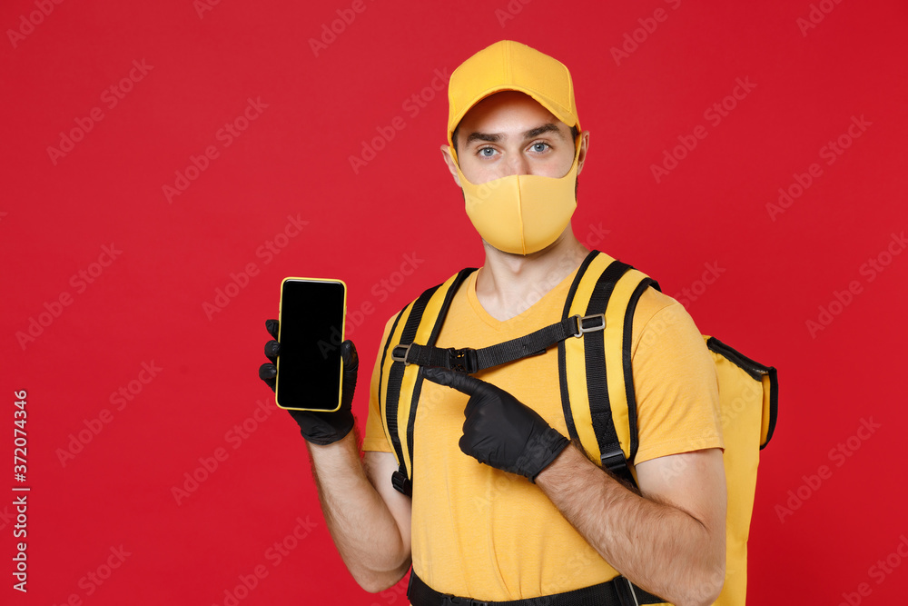 Delivery employee man in yellow cap face mask gloves t-shirt uniform thermal food bag backpack, hold cellphone, work courier service during quarantine covid-19 virus isolated on red background studio. Delivery employee man in yellow cap face mask gloves t-shirt uniform thermal food bag backpack, hold cellphone, work courier service during quarantine covid-19 virus isolated on red background studio.