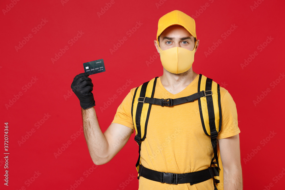 Delivery employee man in yellow cap face mask gloves t-shirt uniform thermal food bag backpack hold credit card work courier service during quarantine covid-19 virus isolated on red background studio.