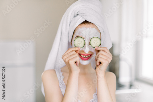 Funny young Caucasian girl with clay mask for face skin and cucumbers on eyes, posing at light home kitchen and having fun, enjoying spa beauty procedures at home