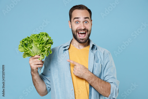 Surprised young bearded man guy 20s wearing casual clothes posing pointing index finger on bunch of fresh greens salad looking camera isolated on pastel blue color wall background studio portrait.