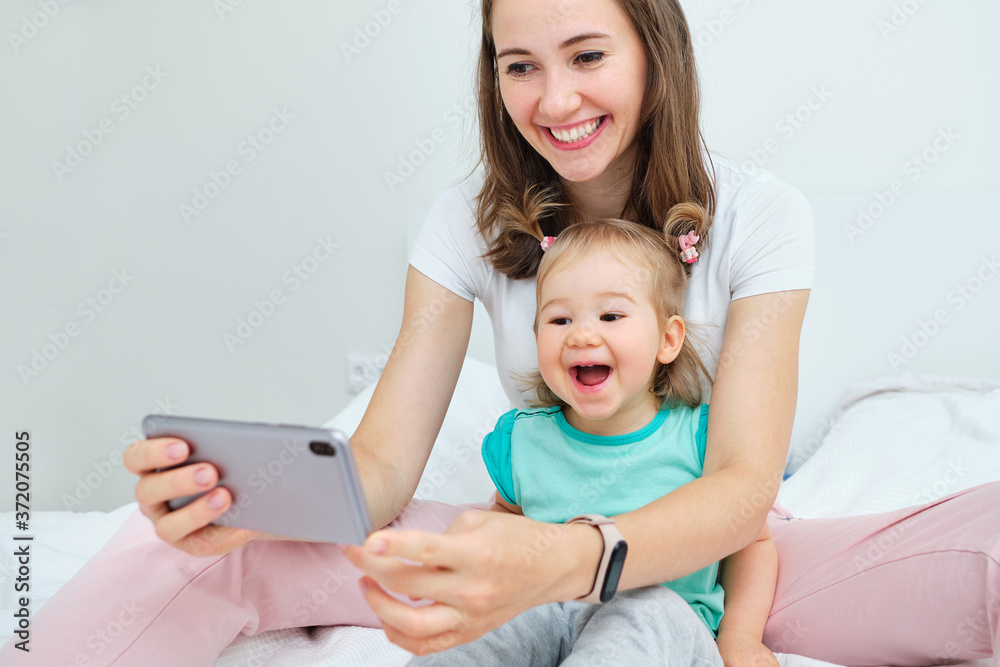 Happy and smiling baby and mom take a photo selfies on their smartphone while sitting on the bed. Happy and smiling baby and mom take a photo selfies on their smartphone while sitting on the bed.