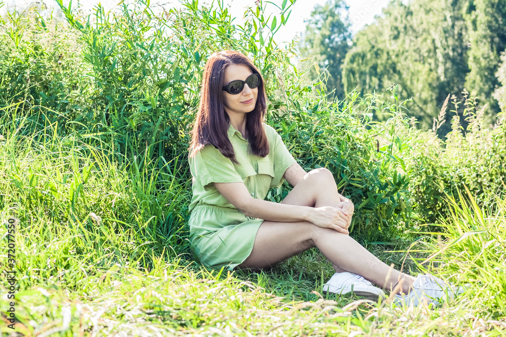 young woman in a pistachio-colored dress sits in tall green grass. Pretty female in sunglasses is enjoying summer nature. Model poses in meadow young woman in a pistachio-colored dress sits in tall green grass. Pretty female in sunglasses is enjoying summer nature. Model poses in meadow